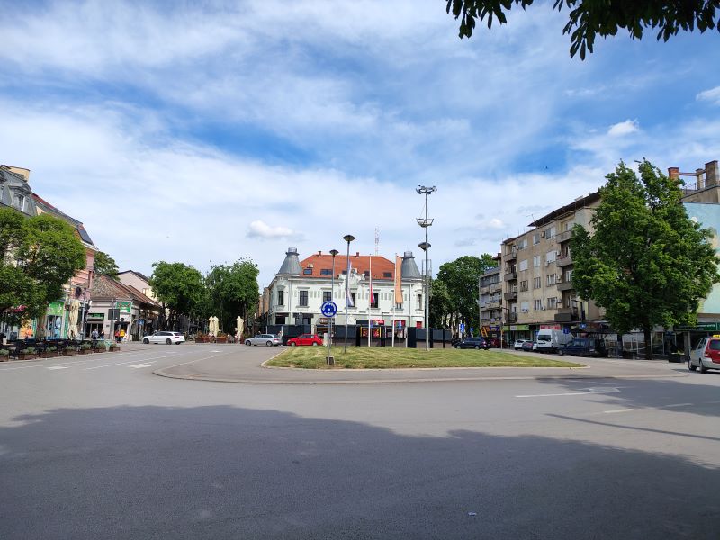 Sunčan pogled na centar grada sa kružnim tokom bez automobila, okruženim zgradama i zelenilom pod vedrim nebom.